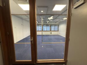 Glass door view into an empty office space with blue carpet at De Ruijterkade.